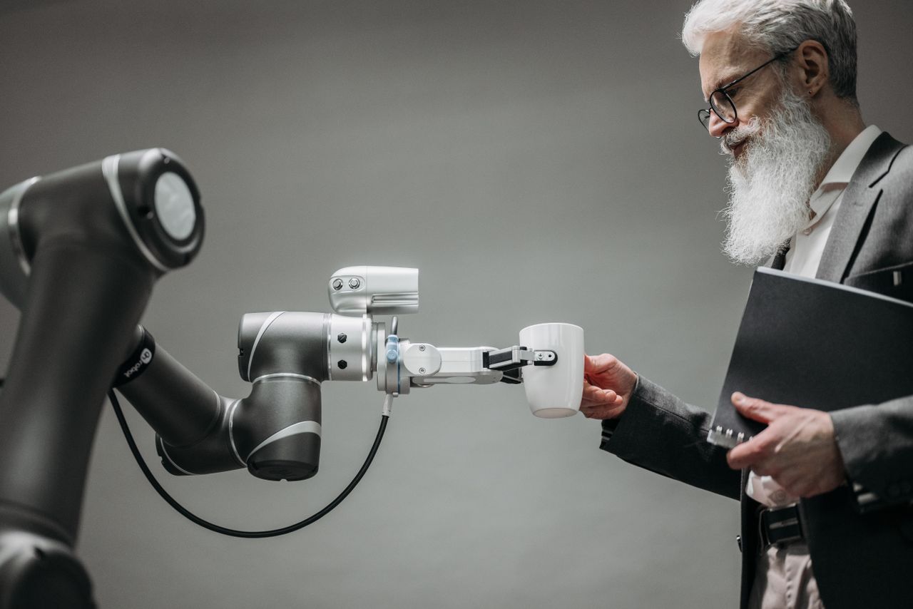 services-01 An elderly man receives a cup from a robotic arm in a modern office setting.